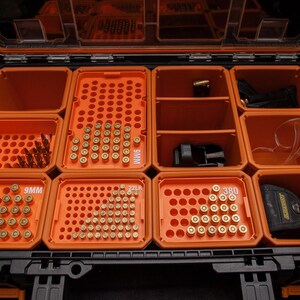 May include: An open, black and orange, divided storage case filled with ammunition and accessories. The case has multiple compartments containing various calibres of bullets, safety glasses, and a holster.