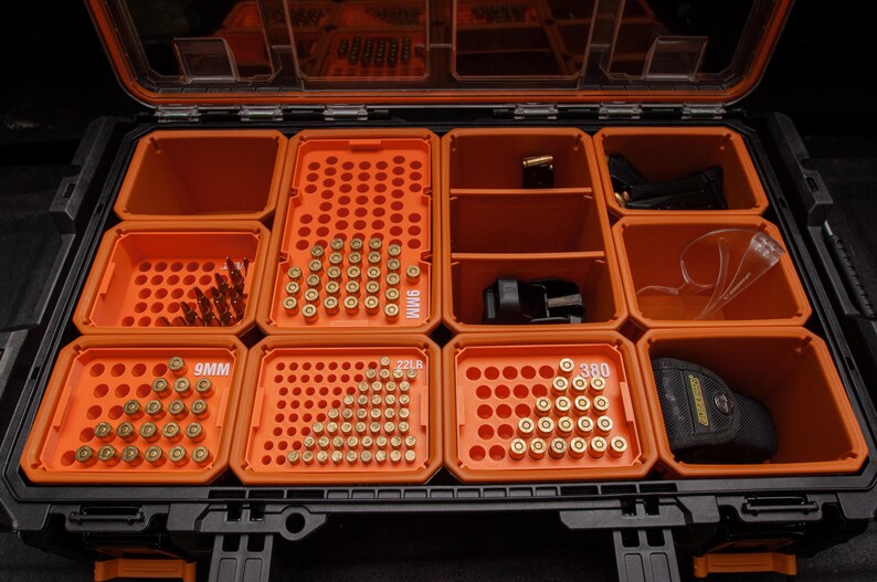 May include: An open, black and orange, divided storage case filled with ammunition and accessories. The case has multiple compartments containing various calibres of bullets, safety glasses, and a holster.