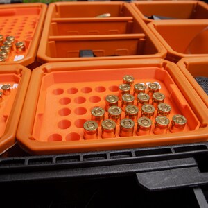 May include: An orange storage case with multiple compartments, each containing brass-coloured ammunition. The case is divided into several sections, each holding a different quantity of bullets. A black, textured base supports the case.