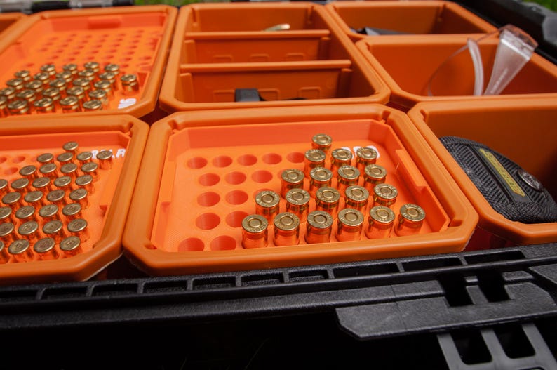 May include: An orange storage case with multiple compartments, each containing brass-coloured ammunition. The case is divided into several sections, each holding a different quantity of bullets. A black, textured base supports the case.
