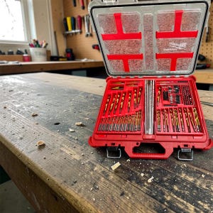 Puede incluir: Un maletín organizador rojo Milwaukee Packout con tapa transparente, que contiene un juego completo de brocas metálicas doradas y brocas escalonadas dispuestas en compartimentos de plástico rojo moldeado. El maletín está abierto.