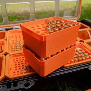 May include: A black and clear plastic storage container with orange trays. Two orange trays are stacked, filled with small, gold-coloured bullets. Other trays contain bullets and small tools.