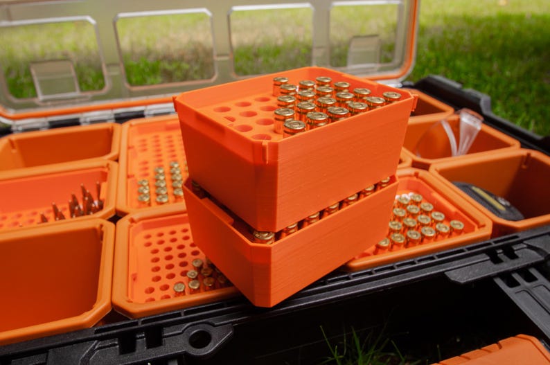 May include: A black and clear plastic storage container with orange trays. Two orange trays are stacked, filled with small, gold-coloured bullets. Other trays contain bullets and small tools.