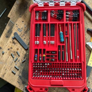May include: A red toolbox filled with various screwdriver bits and accessories. The bits are organized in compartments, with black and red color schemes. A clear lid is visible, and the toolbox is on a wooden surface.