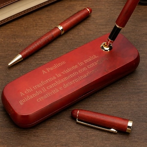 May include: A set of two dark red wooden pens with gold-tone accents and a matching wooden pen stand. One pen is inserted into the stand, which features the engraved Italian text: A Paolino: A chi trasforma la visione in realtà, guidando il cambiamento con coraggio, creatività e determinazione.