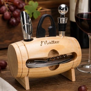May include: A light-coloured wooden wine accessory holder shaped like a barrel, featuring the name Matteo engraved on the front. It contains a metal wine pourer, a black foil cutter, a metal bottle stopper, and a folding corkscrew tool.