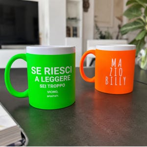 May include: A bright green mug with white text reading SE RIESCI A LEGGERE SEI TROPPO VICINO, SPOSTATI. and a bright orange mug with white text reading MA ZIO BILLY. Both mugs have white interiors and matching coloured handles.