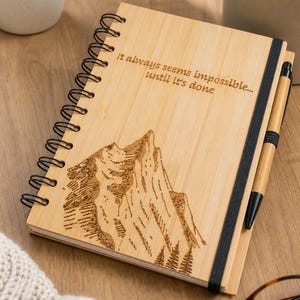Pode incluir: Um caderno de espiral com capa de madeira clara apresentando uma paisagem de montanha gravada a laser e o texto "It always seems impossible... until it's done.". Uma caneta de madeira a condizer é mantida por uma fita elástica preta.