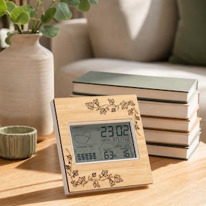 Op de afbeelding: Een vierkant digitaal weerstation met een licht bamboehouten frame voorzien van lasergegraveerde rankpatronen. Het scherm toont de tijd 23:02, een weersverwachtingsicoon, een luchtvochtigheid van 63% en een temperatuur van 16,9 graden Celsius.