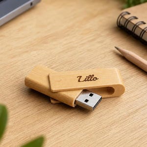 Può includere: Una chiavetta USB in legno chiaro con cappuccio girevole. La parola Lillo è incisa in un carattere corsivo scuro sulla superficie superiore dell'involucro in legno. Il connettore USB in metallo è esposto e sporge dal corpo in legno.