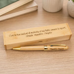 Könnte beinhalten: Ein heller Holzstift und ein passendes rechteckiges Holz-Etui. Der Stift hat goldfarbene Metallakzente und den eingravierten Text "Maestro Angelo". Der Deckel des Etuis ist mit "Con le tue lezioni di motoria ci hai fatto amare lo sport. Grazie maestro Angelo" graviert.