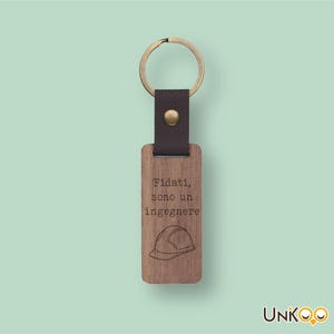 Op de afbeelding: Een rechthoekige houten sleutelhanger met een donkerbruine leren band, een messingkleurige metalen klinknagel en een ronde ring. Het hout is gegraveerd met de tekst "Fidati, sono un ingegnere" boven een lijntekening van een veiligheidshelm.
