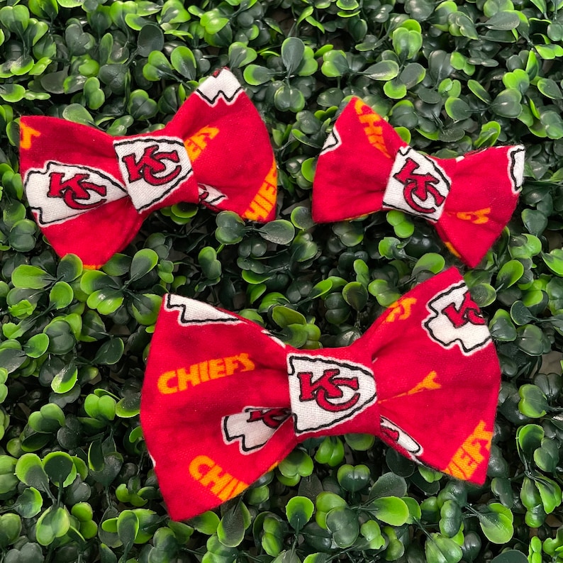 Op de afbeelding: Drie rode strikjes met een wit en geel ontwerp. De strikjes hebben het logo van de Kansas City Chiefs. De strikjes zijn gerangschikt op een bed van groen gebladerte.