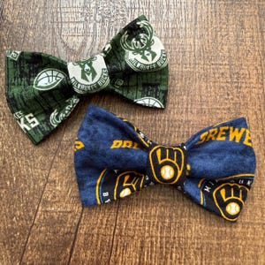 May include: Two bow ties with sports team logos. One is green with a basketball and the words "Milwaukee Bucks". The other is navy blue with a baseball and the word "Brewers". Both are made of fabric.