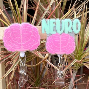 May include: Two pink glitter brain-shaped badge holders with clear retractable reels. The top badge holder has the word "NEURO" written in green glitter on a silver background.