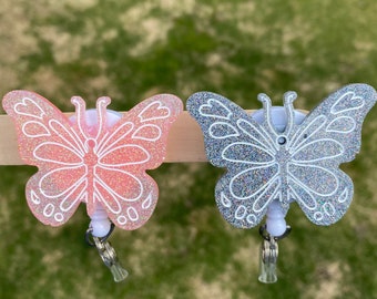 May include: Two glitter butterfly badge reels, one pink and one silver. The reels are attached to a white background.