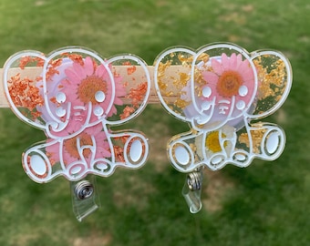 May include: Two white resin elephant shaped badge reels with gold and pink pressed flowers and gold leaf accents.