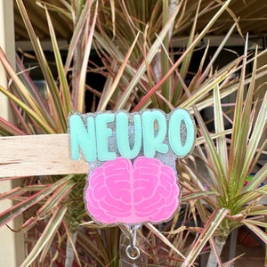 May include: A teal blue badge holder with the word "NEURO" in white glitter lettering. A pink brain graphic is attached to the holder, and a clear retractable badge reel is attached to the bottom.