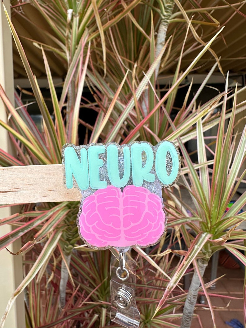 May include: A teal blue badge holder with the word "NEURO" in white glitter lettering. A pink brain graphic is attached to the holder, and a clear retractable badge reel is attached to the bottom.