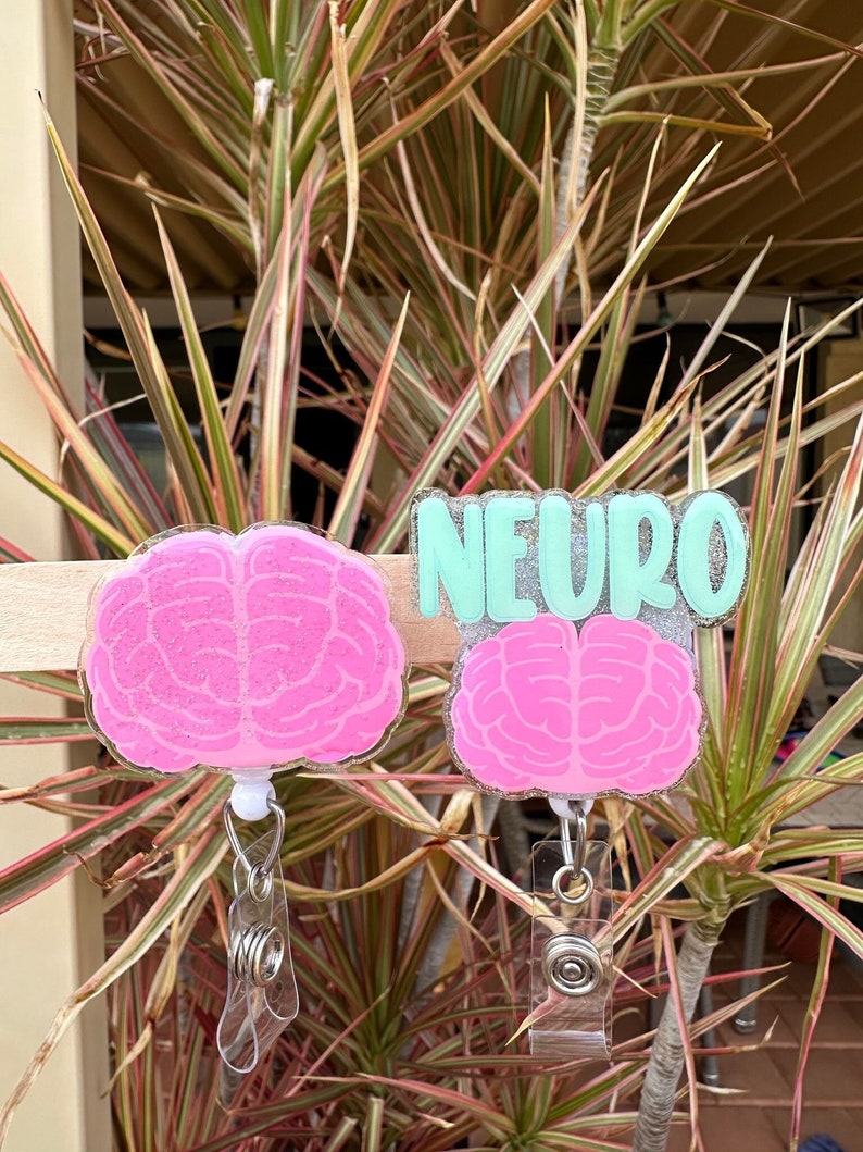 May include: Two pink glitter brain-shaped badge holders with clear retractable reels. The top badge holder has the word "NEURO" written in green glitter on a silver background.
