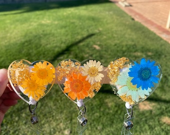 May include: Three heart-shaped resin keychains with different pressed flower designs. The keychains are yellow, orange, and blue with gold flakes. Each keychain has a clear plastic retractable badge holder.