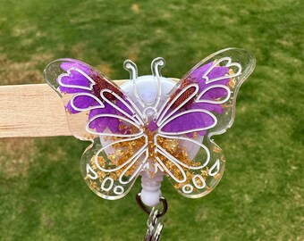 May include: A clear plastic butterfly-shaped badge holder with a purple flower design and gold glitter. The holder has a retractable badge reel and a metal clip.