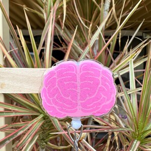 May include: Pink brain-shaped badge holder with glitter. The holder has a white clip and a silver retractable badge reel.