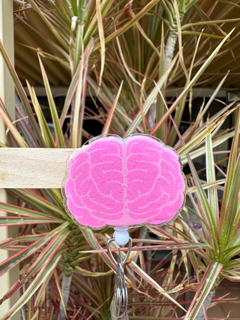 May include: Pink brain-shaped badge holder with glitter. The holder has a white clip and a silver retractable badge reel.