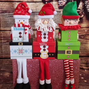 May include: Three Christmas gift boxes with legs. The boxes are decorated with Santa, a snowman, and an elf. The boxes are labelled "Millie", "Thomas", and "Sophie".