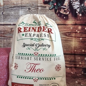 May include: A white canvas Santa sack with red and green text that reads "Reindeer Express Special Delivery Overnight Service For: Theo Checked Twice By: Santa Claus Deliver By: Dec 25 Deliverable. Return To: Santa Claus, North Pole"