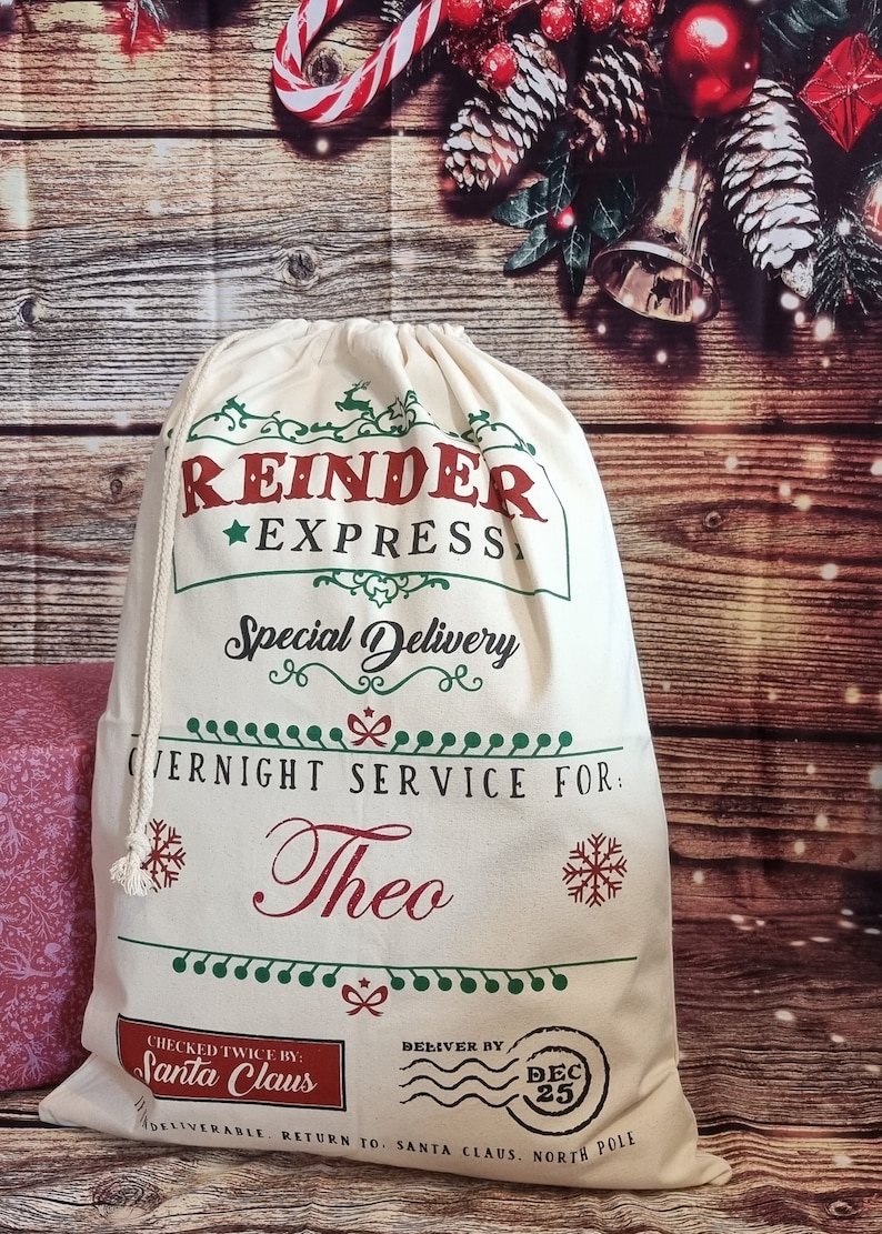 May include: A white canvas Santa sack with red and green text that reads "Reindeer Express Special Delivery Overnight Service For: Theo Checked Twice By: Santa Claus Deliver By: Dec 25 Deliverable. Return To: Santa Claus, North Pole"