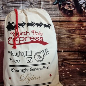 May include: A white canvas Santa sack with red trim and black lettering. The sack says "North Pole Express" and has a check box for "Nice" and a blank box for "Naughty". It also says "Overnight Service For Dylan".