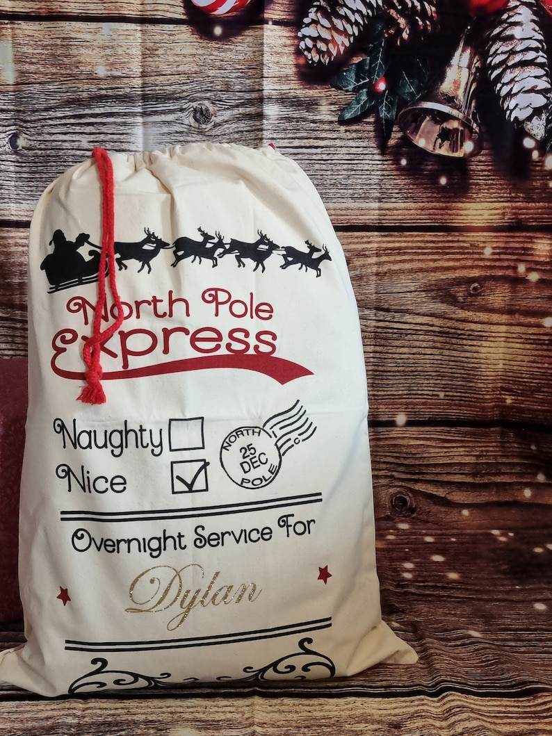 May include: A white canvas Santa sack with red trim and black lettering. The sack says "North Pole Express" and has a check box for "Nice" and a blank box for "Naughty". It also says "Overnight Service For Dylan".