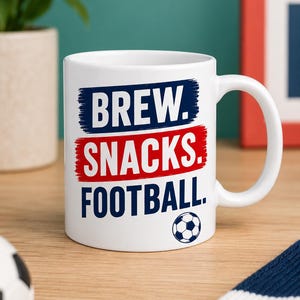 May include: A white ceramic mug featuring the text BREW. in dark blue, SNACKS. in red, and FOOTBALL. in dark blue, with a small black and white football graphic positioned below the text. The mug has a standard handle and a cylindrical shape.