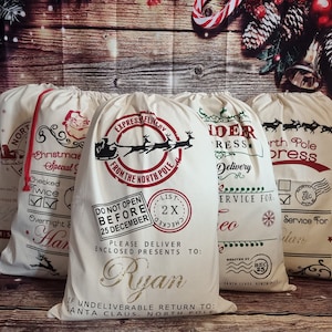 May include: Four white canvas Santa sacks with red and green designs. The sacks have different text, including "Express Delivery from the North Pole", "North Pole Express", and "Checked Twice". One sack has the name "Ryan" written on it.