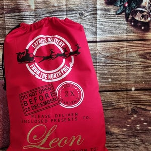 May include: Red Christmas gift bag with a white circle that says "Express Delivery from the North Pole".  The bag also has a white stamp that says "Do Not Open Before 25 December" and a white stamp that says "List Checked 2x".  The bag has a gold glitter name that says "Leon" and the text "Please deliver enclosed presents to:  Undelieverable return to: Santa Claus, North Pole".