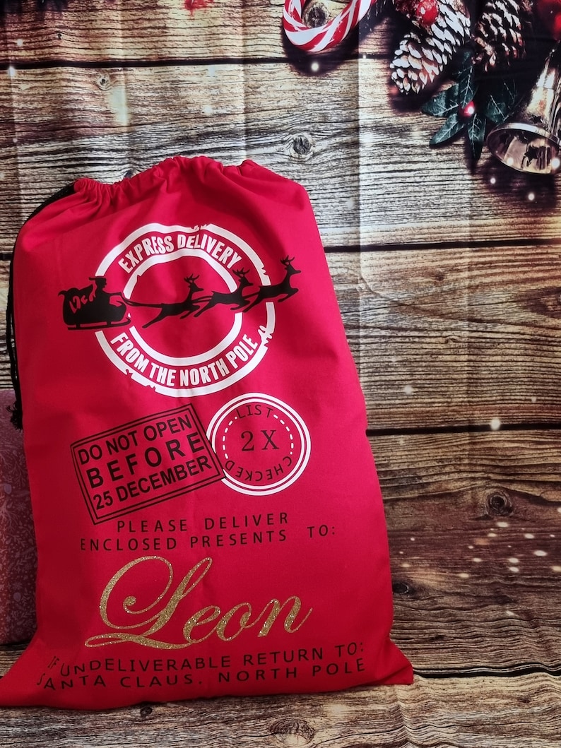 May include: Red Christmas gift bag with a white circle that says "Express Delivery from the North Pole".  The bag also has a white stamp that says "Do Not Open Before 25 December" and a white stamp that says "List Checked 2x".  The bag has a gold glitter name that says "Leon" and the text "Please deliver enclosed presents to:  Undelieverable return to: Santa Claus, North Pole".