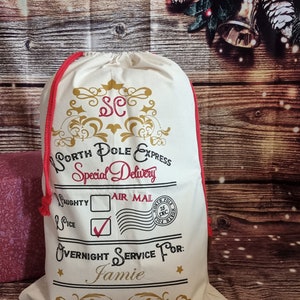 May include: A white canvas Santa sack with red drawstring, gold lettering, and a black and white stamp that says "North Pole Express Special Delivery" and "Air Mail". The sack has a check mark next to "Nice" and a box next to "Naughty". The stamp also says "North Pole 25 Dec". The sack is personalized with the name "Jamie".
