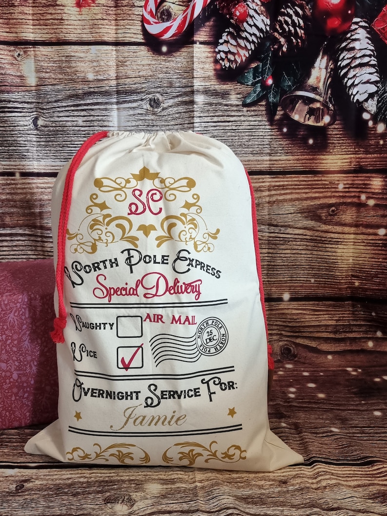 May include: A white canvas Santa sack with red drawstring, gold lettering, and a black and white stamp that says "North Pole Express Special Delivery" and "Air Mail". The sack has a check mark next to "Nice" and a box next to "Naughty". The stamp also says "North Pole 25 Dec". The sack is personalized with the name "Jamie".