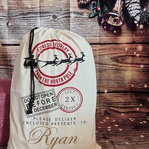 May include: A white canvas Santa sack with red and black lettering. The sack has a drawstring closure and says "Express Delivery from the North Pole". It also has a red stamp that says "Do Not Open Before December" and another red stamp that says "List 2X Checked". The sack is personalized with the name "Ryan".