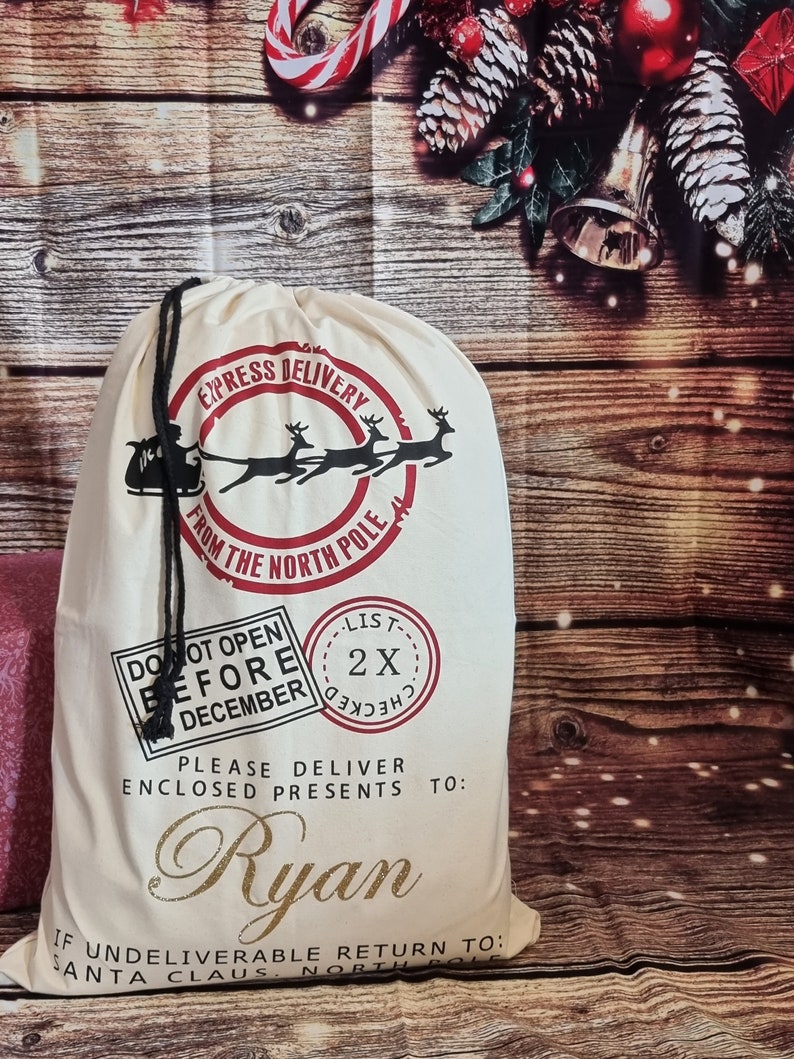 May include: A white canvas Santa sack with red and black lettering. The sack has a drawstring closure and says "Express Delivery from the North Pole". It also has a red stamp that says "Do Not Open Before December" and another red stamp that says "List 2X Checked". The sack is personalized with the name "Ryan".