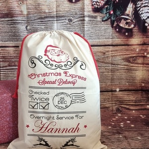 May include: A white canvas Santa sack with red trim and a Santa Claus graphic. The sack has the text "Christmas Express Special Delivery" and "Checked Twice" with check marks. It also has a red and white North Pole stamp with the date "25 DEC". The sack says "Overnight Service For Hannah" with a star on either side of the name.
