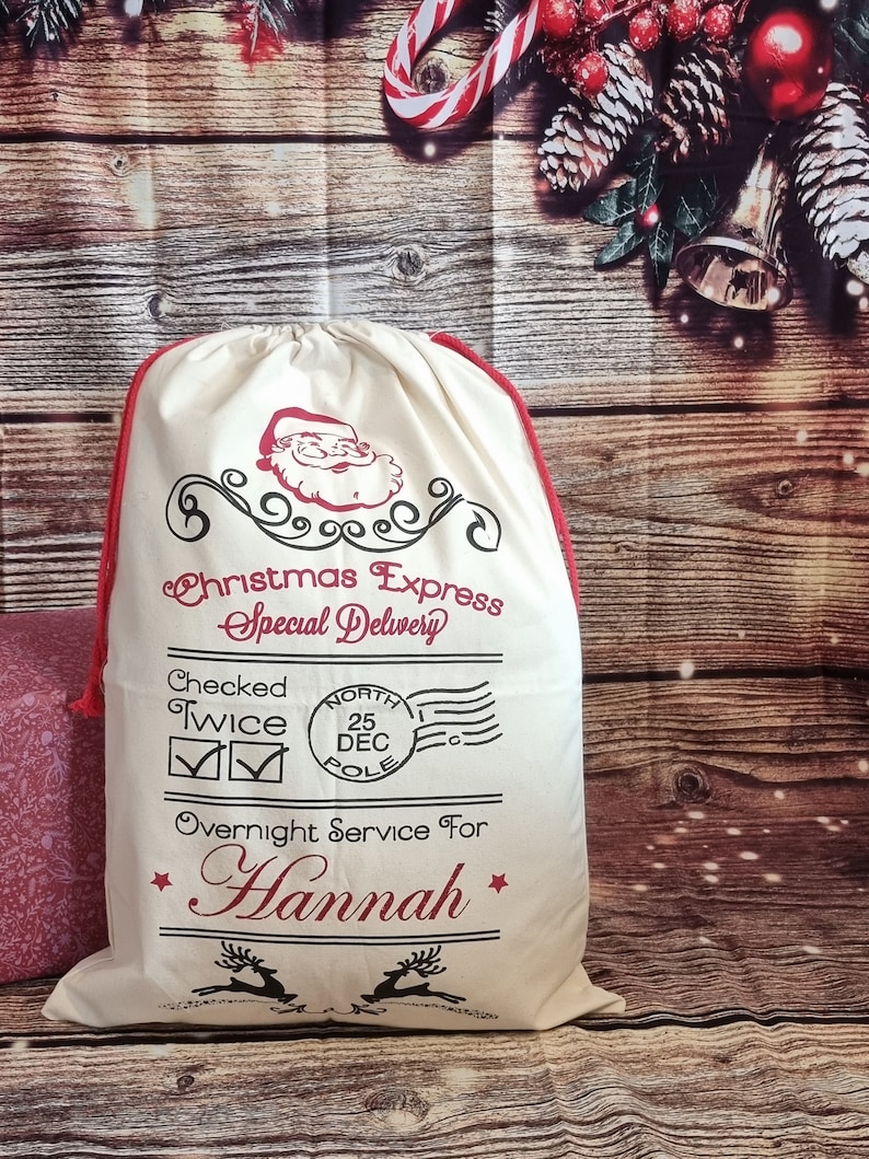 May include: A white canvas Santa sack with red trim and a Santa Claus graphic. The sack has the text "Christmas Express Special Delivery" and "Checked Twice" with check marks. It also has a red and white North Pole stamp with the date "25 DEC". The sack says "Overnight Service For Hannah" with a star on either side of the name.