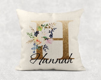 May include: A white pillow with a gold letter H decorated with watercolor flowers and the name "Hannah" written in black cursive.