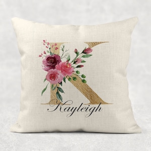 May include: A white cushion featuring a gold letter K adorned with pink and burgundy watercolour flowers. The name Kayleigh is written in cursive beneath the letter.