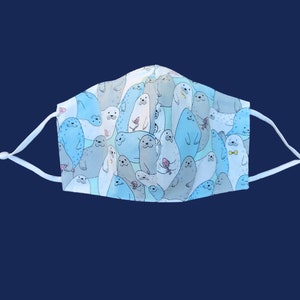 May include: A reusable face mask featuring a repeating pattern of cartoon seals in shades of blue and gray. The seals have various expressions and hold items like ice cream cones. The mask has white adjustable ear loops.