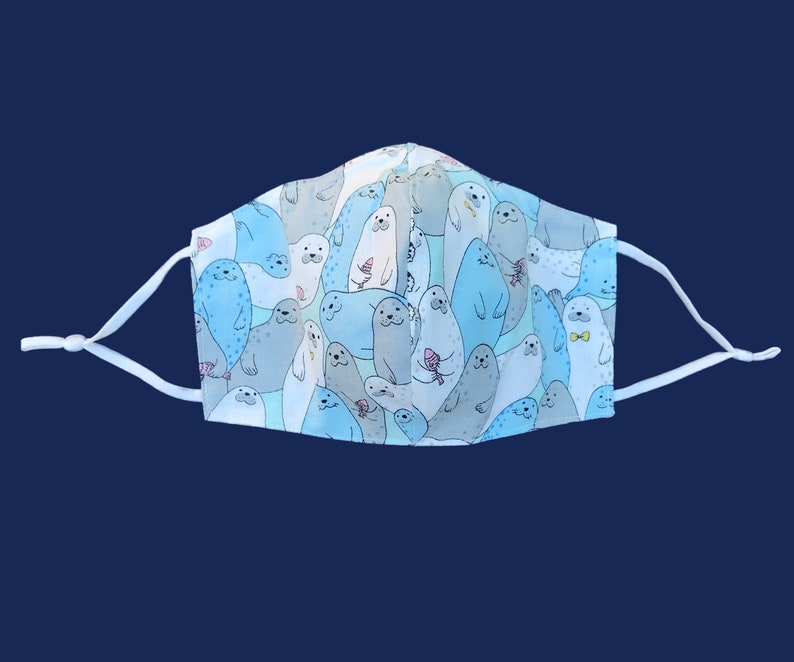May include: A reusable face mask featuring a repeating pattern of cartoon seals in shades of blue and gray. The seals have various expressions and hold items like ice cream cones. The mask has white adjustable ear loops.