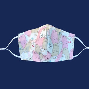 May include: A white face mask with a repeating pattern of cartoon seals in pink, tan, and white. The seals have various expressions and some wear bow ties. The mask has adjustable white ear loops.