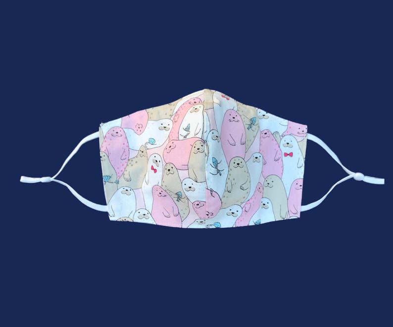 May include: A white face mask with a repeating pattern of cartoon seals in pink, tan, and white. The seals have various expressions and some wear bow ties. The mask has adjustable white ear loops.