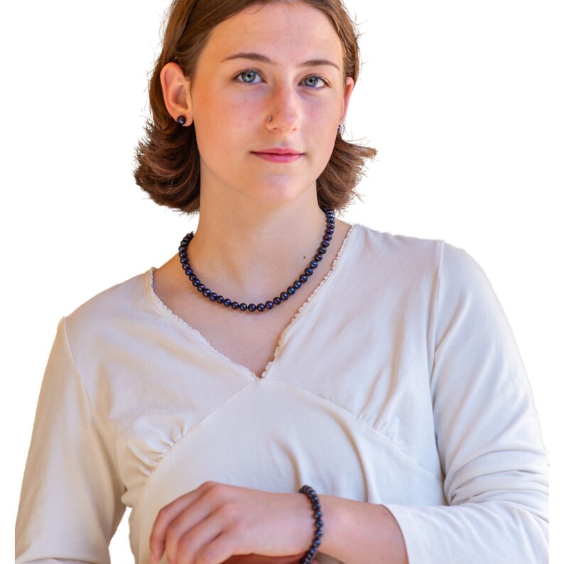 May include: A dark blue pearl necklace, bracelet, and matching stud earrings. The jewelry is displayed on a person wearing a cream-colored top. The pearls have a lustrous sheen, and the set is a classic accessory.